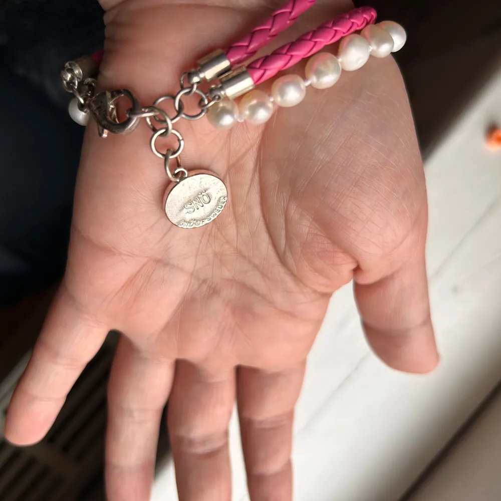 Snyggt armband med en mix av rosa flätat band och vita Sötvattenpärlor . Armbandet har en silverfärgad kedja och ett litet runt hänge. Perfekt för att ge en färgklick till din outfit!. Accessoarer.