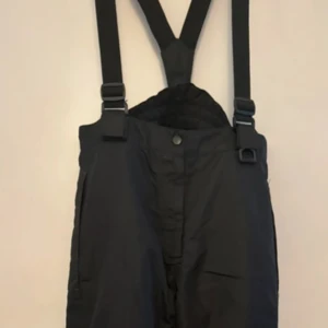 Svarta överdrags med hängslen från Crivit - Säljer ett par svarta överdragsbyxor från Crivit i storlek 16/152. De har praktiska hängslen som går att ta loss och dragkedja framtill. Perfekta för vintern med sitt varma material och snöflingedetalj vid benet. Passar bra för utomhusaktiviteter i kallt väder. Slitage längst ner på benen (se sista bilden) men inget som märks. Kort dragkedja längst ner på benen. 