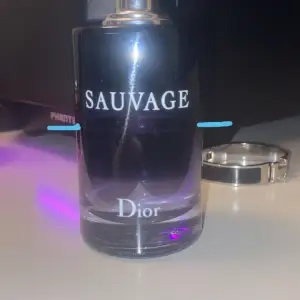 Säljer en flaska Sauvage Eau de Toilette från Dior. Flaskan är elegant med en mörkblå ton och silverdetaljer. Den rymmer 100 ml och har en stilren design med Dior-loggan på toppen. Perfekt för den som vill ha en fräsch och maskulin doft. Passar både till vardags och fest! Ungefär 50ml kvar! INGEN KORK