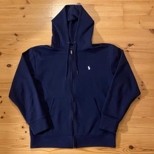 Mörkblå hoodie från Ralph Lauren - Säljer en snygg mörkblå hoodie från Ralph Lauren i mycket bra skick. Den har en dragkedja framtill och en klassisk logga broderad på bröstet. Perfekt för höst och vinter, och den är supermjuk och bekväm. Passar både till vardags och chillkvällar. 💙