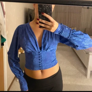 Blå blus med knappar - Snygg blå blus med djup v-ringning och knappar framtill. Den har långa ärmar med en lätt puffig stil och ett blommigt mönster. Perfekt för både vardag och fest!