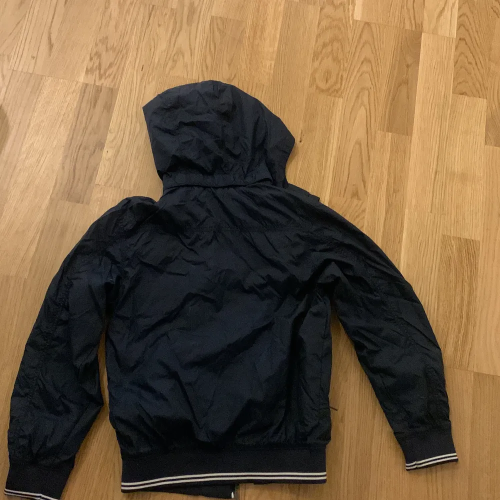 Säljer en mörkblå vindjacka från H&M i storlek 140. Jackan har en huva och dragkedja framtill. Den är fodrad med grått tyg och har ribbade muddar vid ärmslut och nederkant. Perfekt för vår och höst! Aldrig använd, helt ny . Takit.