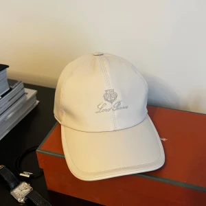 Vit keps från Loro Piana - Snygg vit keps från Loro Piana i ett stilrent och klassiskt utförande. Kepsen har en broderad logga framtill och en elastisk rem baktill för en bekväm passform. Perfekt för både vardag och mer uppklädda tillfällen.