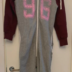 Grå och vinröd onesie med nummer 96 - Supermysig onesie i grått med vinröda ärmar och huva. Den har en stor rosa '96' på framsidan och en praktisk dragkedja som går att dra upp hela vägen. Perfekt för chilliga dagar hemma eller som en cool outfit för en avslappnad dag ute. Passar perfekt för höst och vinter!