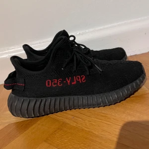 Svarta Adidas Yeezy sneakers - Säljer ett par svarta Adidas Yeezy sneakers i bra skick. De har den klassiska 'SPLY-350' texten i rött på sidan och en bekväm, ribbad sula. Perfekta för både vardag och fest! Skorna har snörning och är i storlek herr 42.
