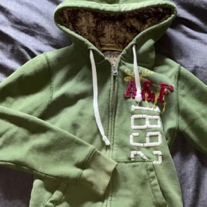 Grön hoodie från Abercrombie & Fitch - Säljer vidare en grön hoodie från Abercrombie & Fitch med dragkedja och huva. Den har en mysig foder inuti och broderade detaljer med 'A&F' och '1892' på framsidan. Perfekt för höst och vinter! Säljer den vidare. 
