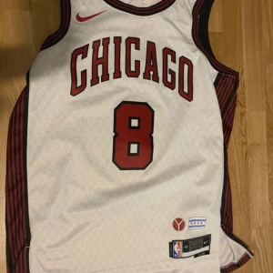 Vit basketlinne från Nike - Säljer en vit basketlinne från Nike med Chicago-tryck och nummer 8. Linnet har svarta och röda detaljer och är perfekt för sportiga aktiviteter. Det är i bra skick och har Dri-FIT-teknologi för att hålla dig sval och torr. Perfekt för basketträning eller som en cool vardagslook! 🏀