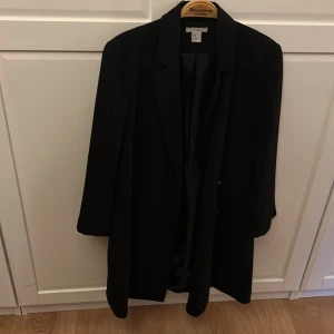 Lång kavaj från H&M - Säljer en stilren svart kavaj från H&M i storlek 40.  Går en bit ner på låret. Gömd knäppning. Perfekt för både vardag och fest. Kavajen har en klassisk design med långa ärmar och en elegant passform. Den är i mycket bra skick och redo för nya äventyr. Passar perfekt till höst- och vårgarderoben!