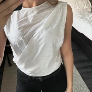 Vit ärmlös topp från Zara - Säljer en stilren vit ärmlös topp från Zara i storlek S. Toppen har en enkel och elegant design med en draperad effekt framtill. Perfekt för både vardag och festliga tillfällen. Passar bra till jeans eller kjol för en fin look. 