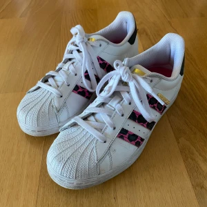 Adidas Superstar med rosa och svarta detaljer - Säljer ett par vita Adidas Superstar i storlek 39 1/3 med rosa och svarta leopardmönster på de tre ränderna. Skorna har snörning och är i bra skick! Den ena skon har lossnat lite där fram nedanför snöret som syns på första bilden men det är inget man tänker på🩷