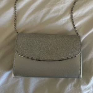 Silverfärgad glittrig kuvertväska - Snygg silverfärgad kuvertväska med glittrig framsida och kedjeaxelrem som går att ta av. Perfekt till balen!❤️  