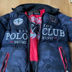 Svart pufferjacka från Santoz Polo Club - Säljer en snygg svart pufferjacka från Santoz Polo Club. Jackan har ett coolt camouflagemönster och stora logotryck både fram och bak. Den är långärmad med dragkedja och har en röd insida som ger en schysst kontrast. Perfekt för höst och vinter!