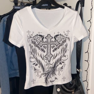 Vit t-shirt med kors och vingar från Romwe - Säljer en ascool vit t-shirt från Romwe med ett stort tryck av ett kors och vingar på framsidan. T-shirten har en v-ringad hals och korta ärmar. Den har skönt material och inte sånt ”konstigt” material som det kan vara från romwe ibland. Använd 1 gång. Säljer då det inte är min stil💕