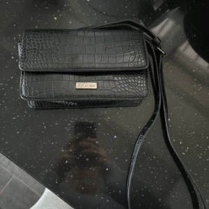 Svart axelväska med krokodilmönster - Snygg svart axelväska i skinn med krokodilmönster. Väskan har en elegant design med en praktisk dragkedja inuti och en justerbar axelrem. Perfekt för både vardag och fest!