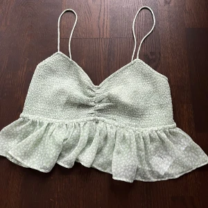 Mint grön blommig topp med volang - Söt grön blommig topp med smala axelband och volangdetalj. Toppen har en smockad design med snörning i ryggen för en justerbar passform. Perfekt för sommaren 💚
