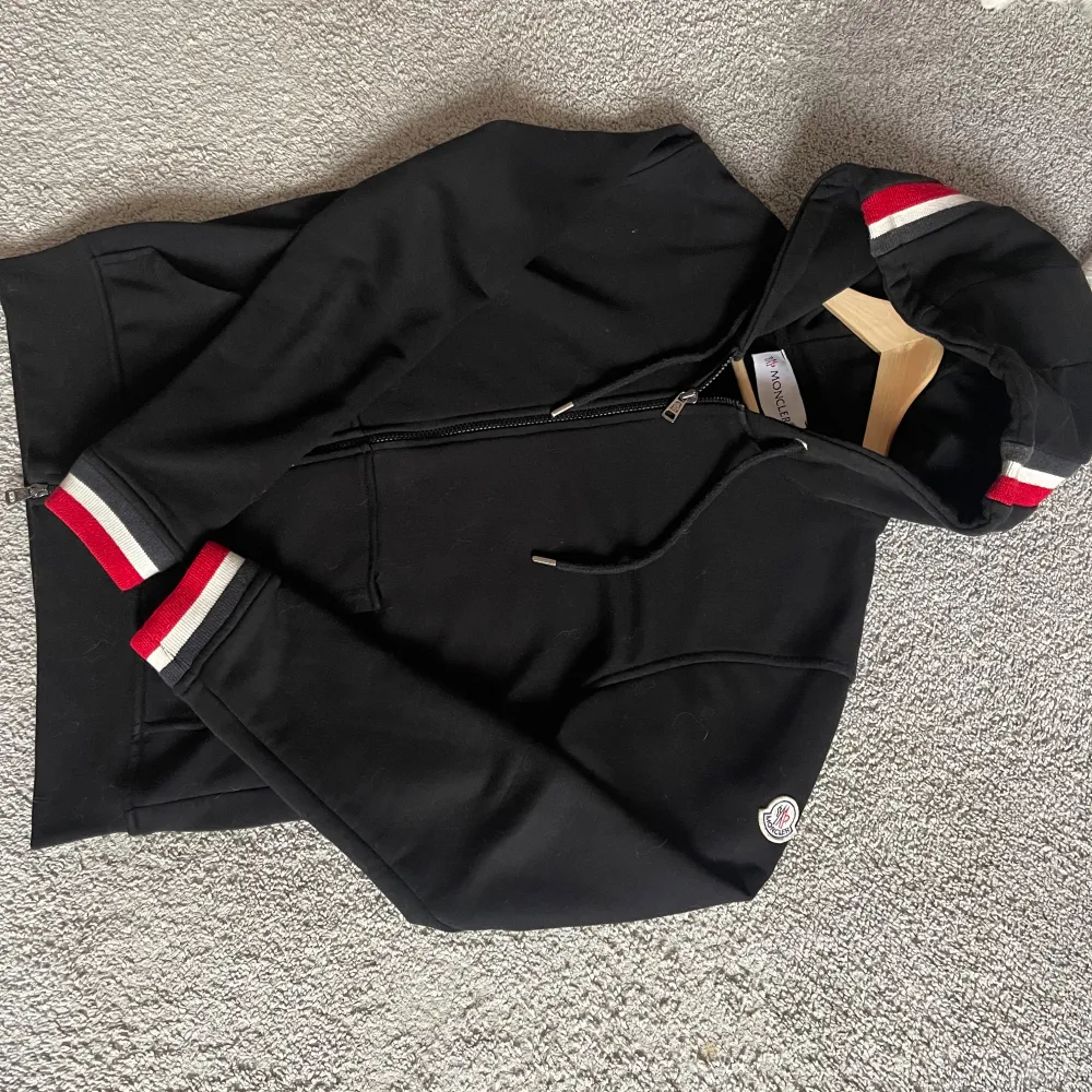 Säljer en snygg svart hoodie från Moncler med dragkedja. Den har coola röda och vita ränder på ärmsluten och luvan. Perfekt för kyliga dagar och att chilla i. Passar både till vardags och lite mer avslappnade tillfällen. 🖤. Tröjor & Koftor.