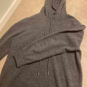 Grå hoodie i kashmir - Mysig grå hoodie i kashmir, perfekt för kyliga dagar. Den har en klassisk design med dragsko i huvan och ribbade muddar vid ärmsluten. Supermjuk och bekväm att ha på sig, passar både till vardags och för mysiga hemmakvällar.