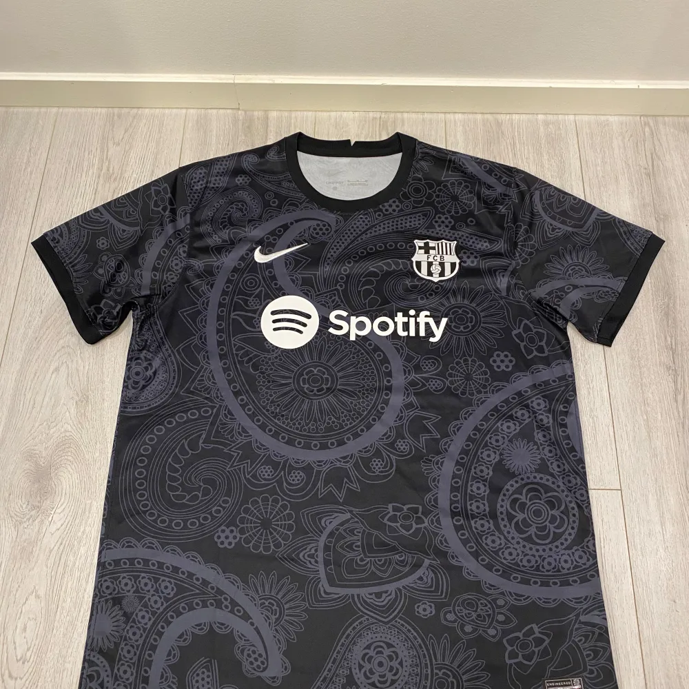 Säljer en snygg svart fotbollströja från Nike med ett coolt paisleymönster. Tröjan har korta ärmar och är nyskick. Den har FCB-loggan och Spotify som sponsortryck på framsidan. Perfekt för fotbollsfans som vill ha något unikt! . T-shirts.