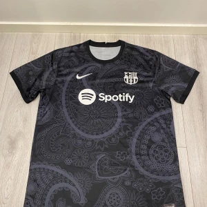 Koncept Nike Barcelona Fotbollströja  - Säljer en snygg svart fotbollströja från Nike med ett coolt paisleymönster. Tröjan har korta ärmar och är nyskick. Den har FCB-loggan och Spotify som sponsortryck på framsidan. Perfekt för fotbollsfans som vill ha något unikt! 