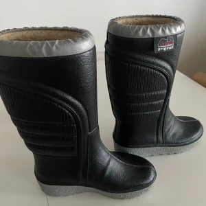 Svarta powerboots med foder - Säljer ett par svarta vinterstövlar i syntetmaterial med varmt foder. Perfekta för kalla vinterdagar. De har en robust design med en grå kant upptill. Använda men i bra skick, redo för nya äventyr i snön!