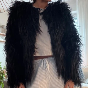 Svart pälsjacka - Säljer en supercool svart pälsjacka i syntetmaterial. Den är perfekt för att ge din outfit en edgy touch. Jackan har långa ärmar och en fluffig look som är både trendig och mysig. Perfekt för höst och vinter, eller varför inte till en fest? Ena fästet sitter lite löst men går superenkelt att sy fast