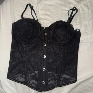 Svart spetskorsett med snörning - Snygg svart korsett i spets med justerbara axelband och snörning bak. Den har hakknäppning fram och ger en elegant look. Perfekt för festliga tillfällen eller en utekväll. Korsetten är i mycket bra skick och har en stilren design.