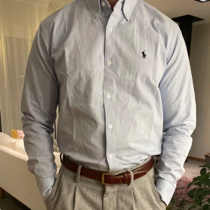 Polo Ralph lauren skjorta  - En klassisk blå skjorta från Polo Ralph Lauren, tillverkad i mjukt bomullstyg för optimal komfort och stil. Med button-down-krage, knäppning framtill och den ikoniska broderade logotypen på bröstet är den både elegant och mångsidig – perfekt för både vardag och fest.