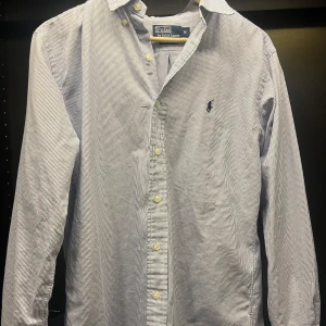 Randig skjorta från Polo Ralph Lauren - Säljer en klassisk blå- och vitrandig skjorta från Polo Ralph Lauren i storlek M. Skjortan har långa ärmar och knappar framtill. Perfekt för både vardag och festliga tillfällen. Den är i mycket bra skick och har en stilren design med broderad logga på bröstet.