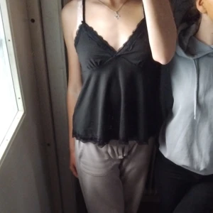 Linne  - Säljer en svart ärmlös topp med smala axelband och spetsdetaljer vid halsringningen. Toppen har en lös passform och är perfekt för sommaren. Den är både stilren och bekväm, vilket gör den idealisk för både vardag och fest.