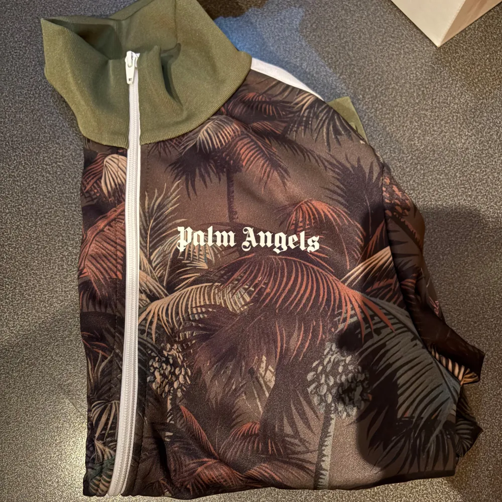 Säljer en snygg jacka från Palm Angels i storlek M. Den har ett coolt mönster med palmer och är i en mörk färgskala. Jackan har dragkedja och är långärmad, perfekt för höst och vår. Materialet är polyester vilket gör den lätt och bekväm. Passar både till vardags och fest!. Tröjor & Koftor.