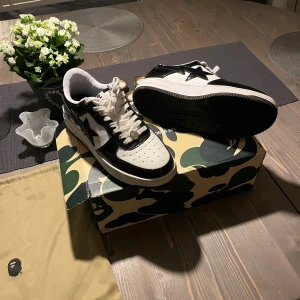 Svartvita sneakers från Bape top skick - Säljer ett par snygga svartvita sneakers från Bape i mycket bra skick. Skorna har en cool stjärndetalj på sidan och kommer med originalkartong i camouflage. Perfekta för både vardag och fest! 🖤🤍