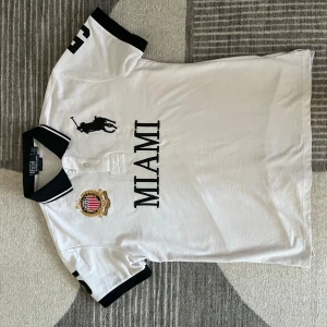 Vit pikétröja från Ralph Lauren - Säljer en snygg vit pikétröja från Ralph Lauren med svart krage och ärmslut. Den har en broderad logga och texten 'MIAMI' på framsidan. Perfekt för en casual look eller en dag på stan. Tröjan är i mycket bra skick och redo för nya äventyr!