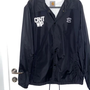 Svart jacka från Carhartt - Säljer en svart Carhartt jacka i nylon med tryck fram och stort logotryck på ryggen. Jackan har knappar framtill och en klassisk krage. Perfekt för vår och höst! 🖤