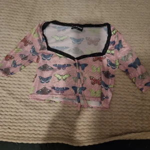Rosa fjärilsmönstrad topp från New Girl Order - Säljer en superfin rosa topp från New Girl Order med färgglada fjärilar över hela tyget. Toppen har trekvartsärmar och en söt knytning framtill. Den är perfekt för vår och sommar och har en lätt och luftig känsla. Passar perfekt till både jeans och kjol!