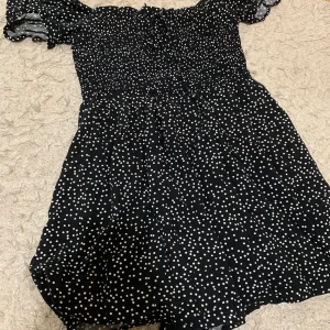Svart prickig klänning från FB Sister - Säljer en superfin svart klänning med vita prickar från Sister. Den har en smockad överdel och korta ärmar med volangdetaljer. Perfekt för sommarens alla fester! 🖤