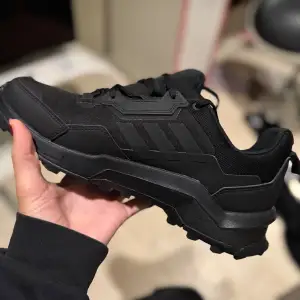 Säljer ett par svarta sneakers från Adidas. De har en robust sula och klassisk snörning, perfekt för att ha utomhus när det är dåligt väder. Skorna är i nytt skick och har en stilren design med de ikoniska tre ränderna på sidan. Det är ett par terrex skor från adidas.