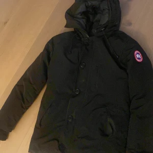 Svart dunjacka från Canada Goose - Säljer en svart dunjacka från Canada Goose i  bra skick. Använt ett par gånger. Jackan har en huva och stängs med både dragkedja och knappar. Perfekt för kalla vinterdagar med sin varma och skyddande design. Märkeslogga på ärmen ger en snygg detalj. Passar perfekt för vintern❄️ PÄLS INGÅR! 