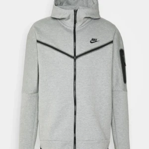 Grå Nike tech  - Säljer en grå hoodie från Nike i mycket bra skick. Den har en dragkedja framtill och en cool design med svarta detaljer. Perfekt för höst och vår, och funkar lika bra till träning som till vardags. Stor ficka på ärmen för extra stilpoäng!