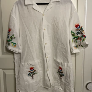 Vit linne skjorta från Zara stl L med broderade blommor - Vit linne skjorta (kortärmad) från Zara i stl L. Skjortan är i 100% linne och är aldrig använd. Skjortan har broderade blommor på sig vilket gör den speciell och lyxig.