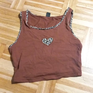 Brun croppad linnetopp med leoparddetaljer - Säljer en brun croppad linnetopp med leopardmönstrade kanter och ett hjärta på bröstet. Perfekt för en avslappnad stil med en touch av vildhet. Toppen är ärmlös och passar bra till både jeans och kjol.