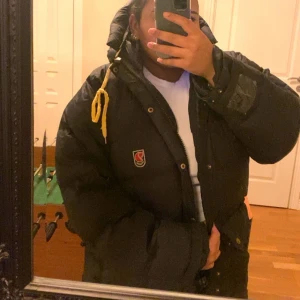 Svart dunjacka från Fjällräven L - Säljer en svart dunjacka från Fjällräven i ett okej skick. Den är använd i två år och har skada på högra armen längst ned. Dragkedjan är också sönder fast det går att laga.☺️