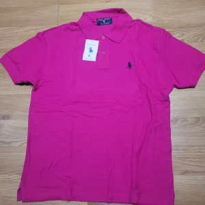 Rosa pikétröja från Polo Sport - Säljer en snygg rosa pikétröja från Polo Sport i 100% bomull. Tröjan har en klassisk krage med knappar och en broderad logga på bröstet. Perfekt för en avslappnad men stilren look. Passar bra till både vardag och fest!