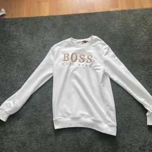 Vit sweatshirt från Hugo Boss - Säljer en stilren vit sweatshirt från Hugo Boss i storlek L. Tröjan har ett snyggt broderat BOSS-märke i guld på framsidan. Perfekt för en avslappnad men ändå trendig look. Passar bra till både jeans och joggers. 🕶️