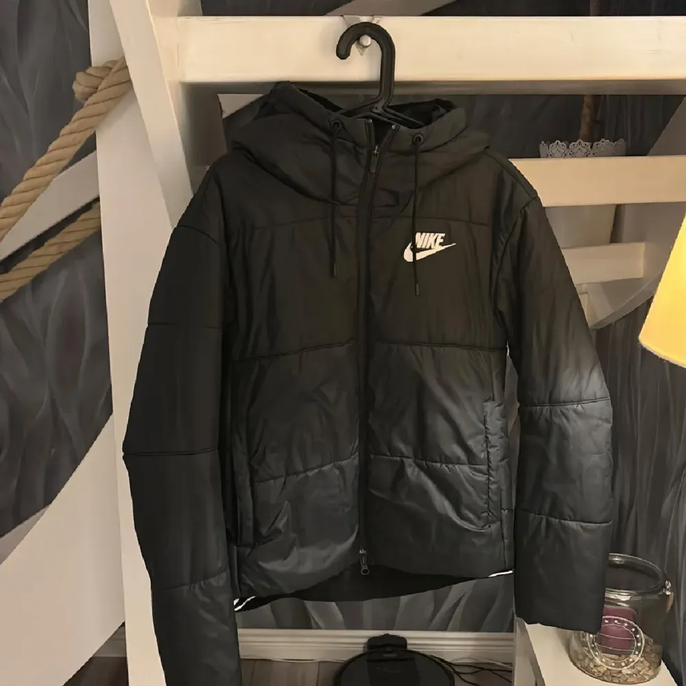 Säljer en svart pufferjacka från Nike i mycket bra skick. Jackan har en huva med dragsko och en dragkedja framtill. Perfekt för kyliga dagar och har en stilren design med Nike-loggan på bröstet. Passar både till vardags och sportiga aktiviteter.. Takit.