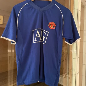 Blå Manchester United fotbollströja - Säljer en blå Manchester United fotbollströja med korta ärmar och vit kant. Den har ett tryck av 'Ronaldo' och nummer 7 på baksidan. Perfekt för fans av laget och spelaren! Tröjan är i bra skick.