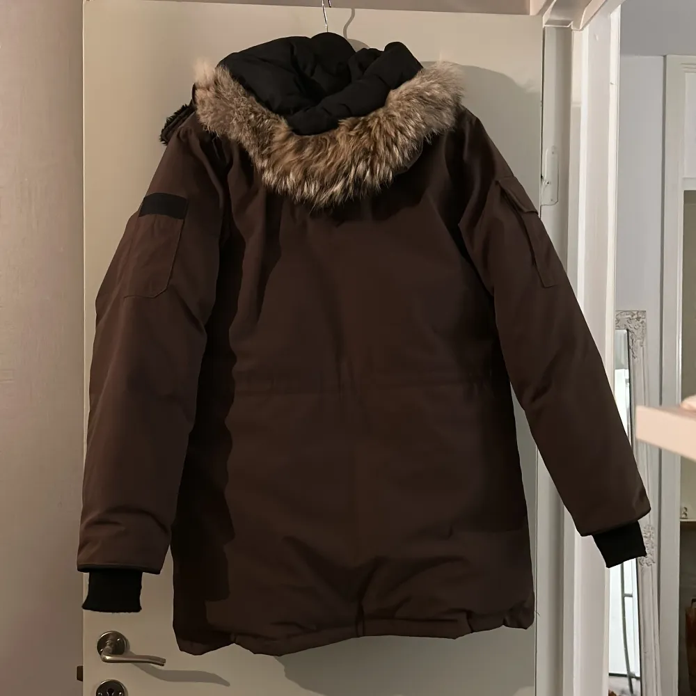 Säljer en brun parkajacka från Canada Goose i mycket bra skick. Jackan har en varm pälsfodrad huva och flera praktiska fickor. Perfekt för kalla vinterdagar. Den är långärmad och har en klassisk design med Canada Goose-logga på bröstet.. Jackor.