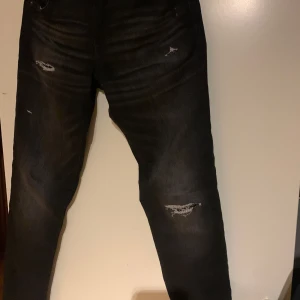 Svarta jeans från Jack & Jones - Säljer ett par svarta jeans från Jack & Jones i bra skick och grå tvätt. De har en snygg slitning på framsidan som ger en cool look. Perfekta för både vardag och fest! 🖤