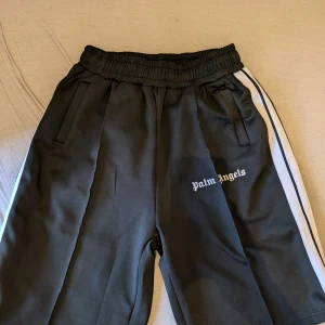 Svarta shorts från Palm Angels - Säljer ett par svarta shorts från Palm Angels i mycket bra skick. De har en elastisk midja och vita ränder längs sidorna. Perfekta för en avslappnad stil eller träning. Passar bra till både vardag och sportiga aktiviteter.