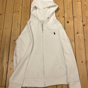 Vit huvtröja från Polo Ralph Lauren - Säljer en snygg vit huvtröja från Polo Ralph Lauren. Den har en dragkedja framtill och en liten broderad logga på bröstet. Perfekt för en avslappnad stil och passar till alla tillfällen. Tröjan har långa ärmar och en skön huva. Perfekt för höst och vår!