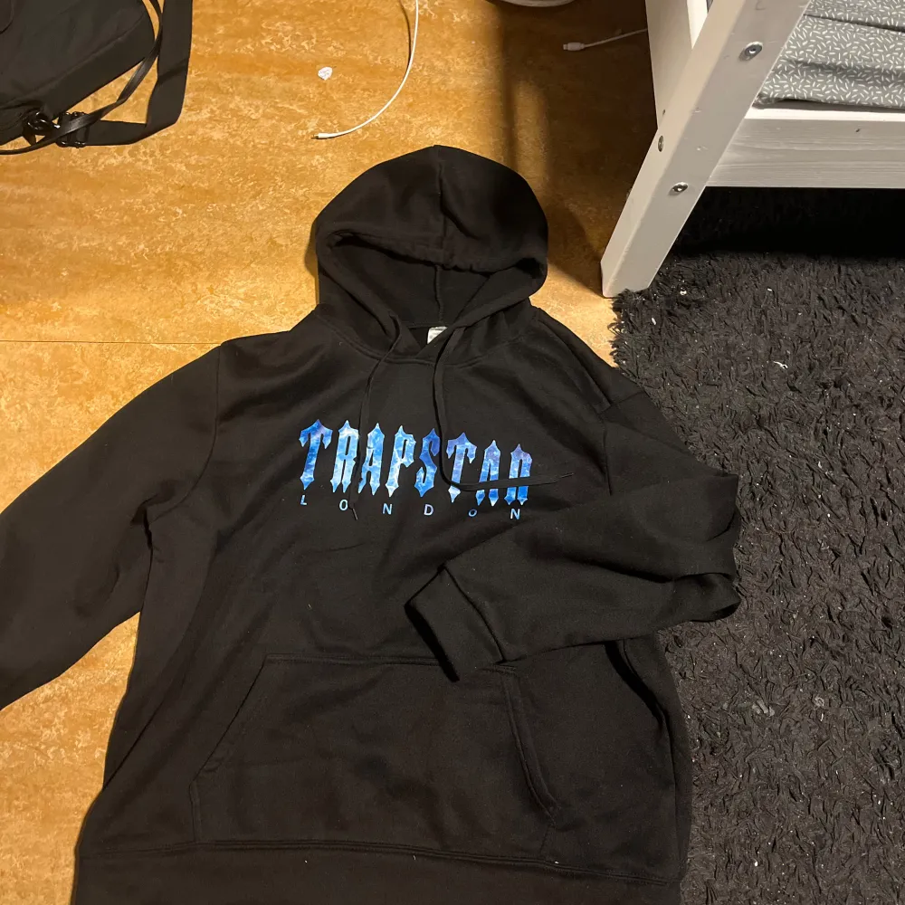 Säljer en svart hoodie från Trapstar med blå text på framsidan. Den har en klassisk känguruficka och en bekväm huva. Perfekt för en avslappnad stil eller streetwear-look. Passar bra till både jeans och joggers.. Tröjor & Koftor.